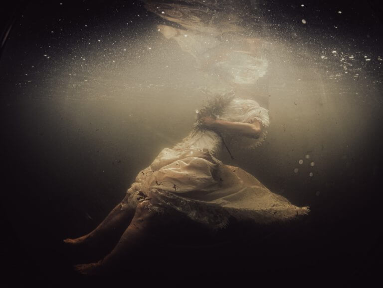 Murky underwater image of female figure, her dress billowing back from the knees, face unseen. This drowned female will need first aid.