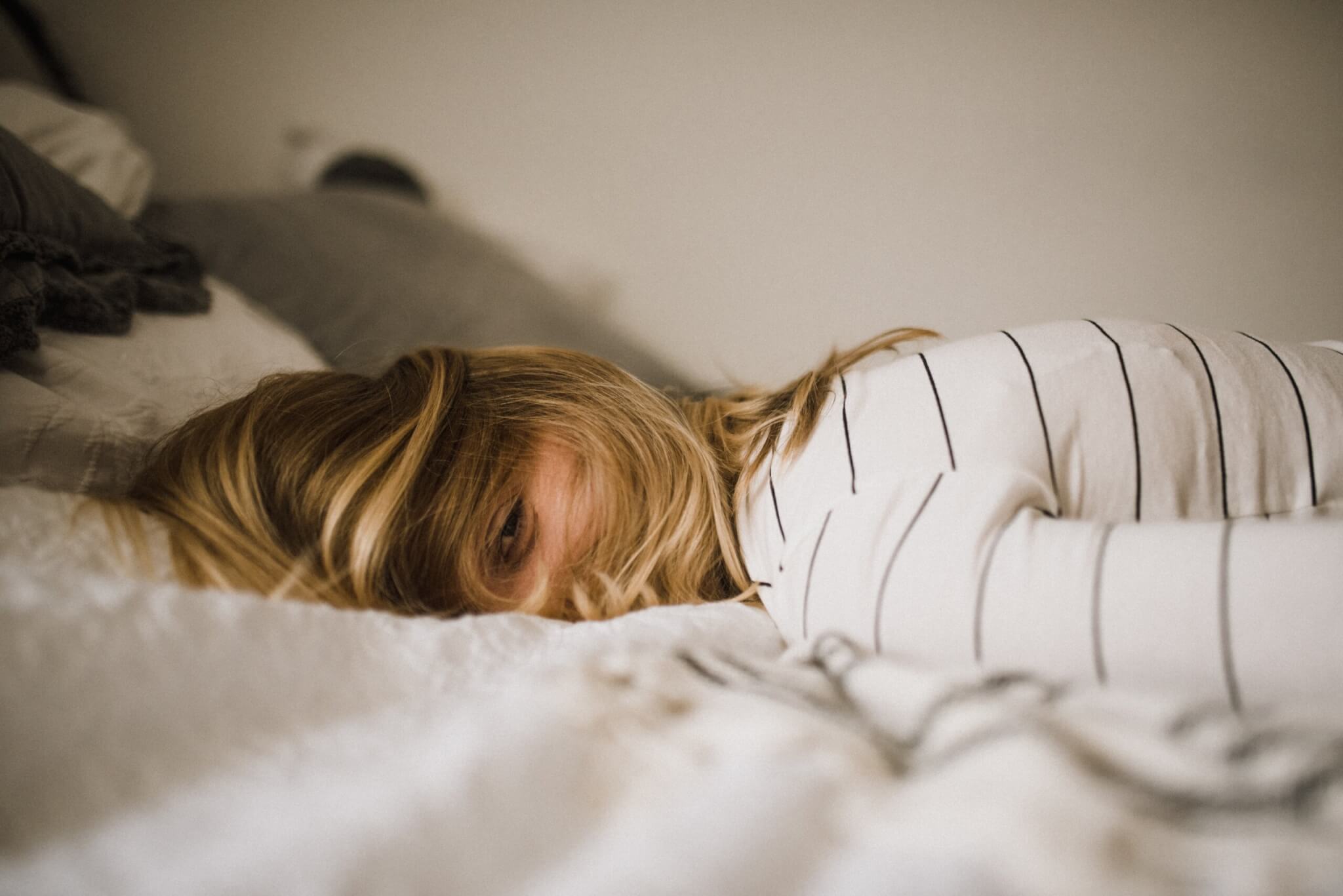 A blond girl lies face down on a bed, eyes mostly closed, hair sprawled over her face. She seems exhausted. 
