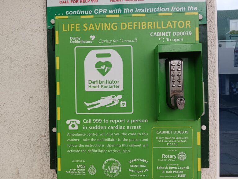 Green housing to a community AED with a number lock. It reads: "Life Saving Defibrillator".