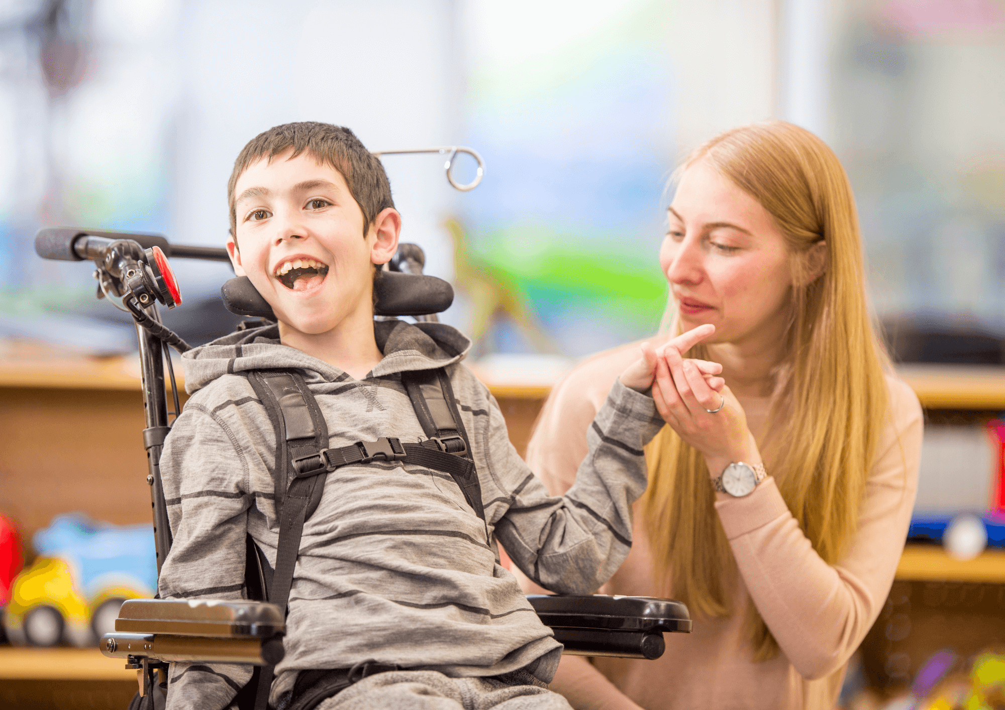 A boy strapped into a wheelchair with supporting headrest smiles with an adult close by in support.